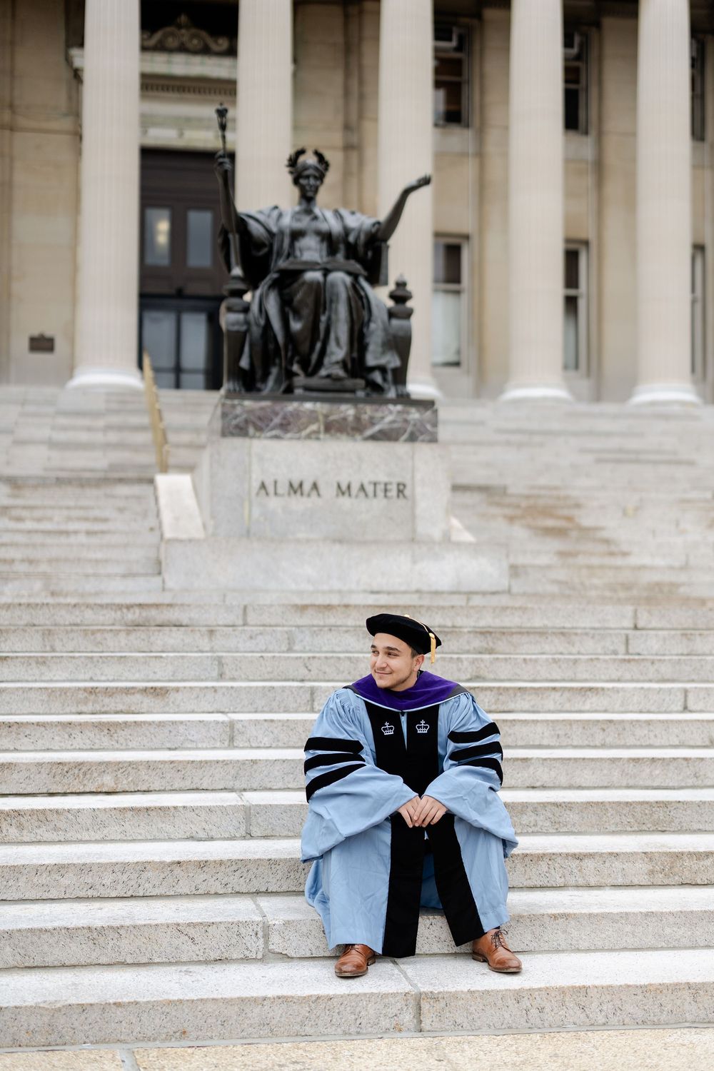 Graduate in academic regalia sits on steps beneath Alma Mater statue at Columbia University.