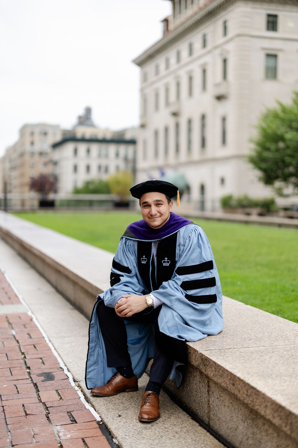 Graduate in academic regalia poses on campus steps in light blue graduation gown and cap.
