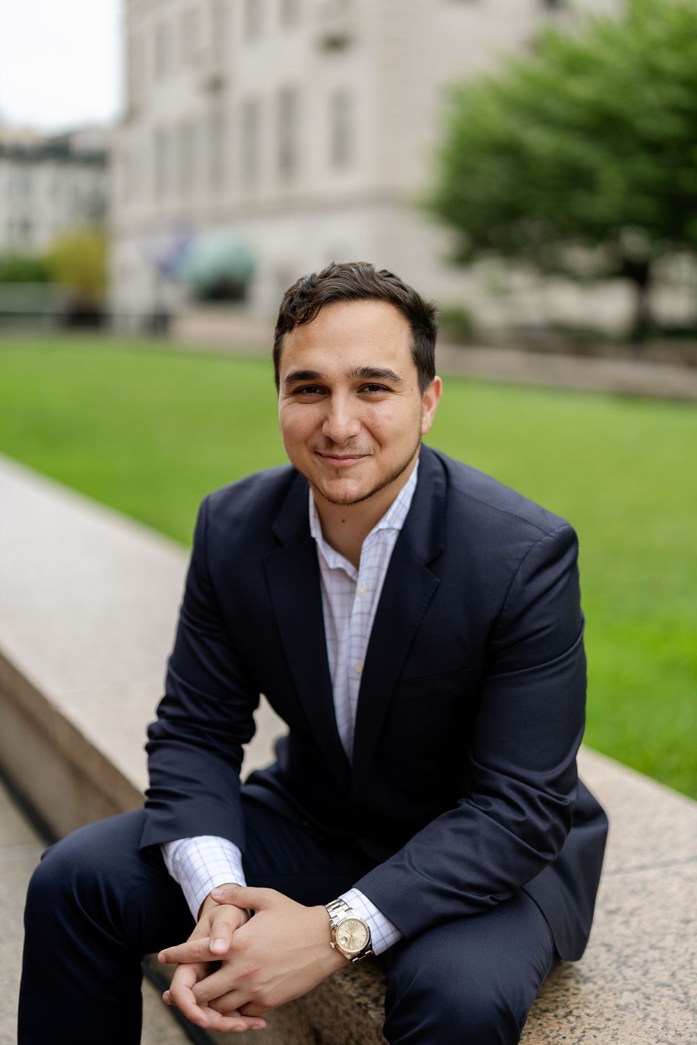 Professional in a navy suit sits on stone ledge outdoors with architectural backdrop.