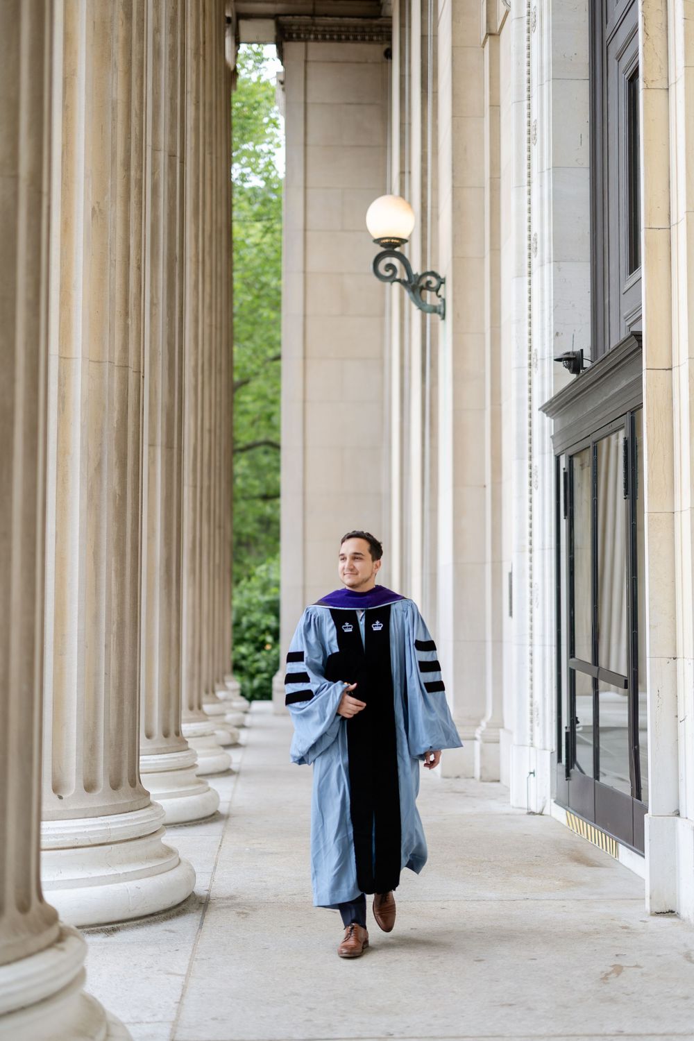 Graduate in academic regalia walks through classical stone columns during commencement ceremony.
