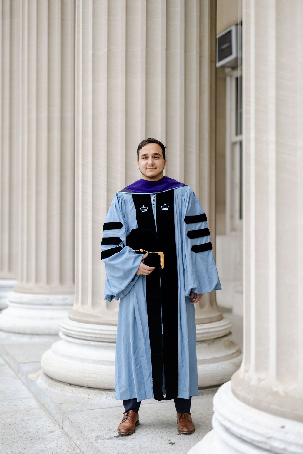 Graduate in academic regalia poses in front of classical stone columns at a university.