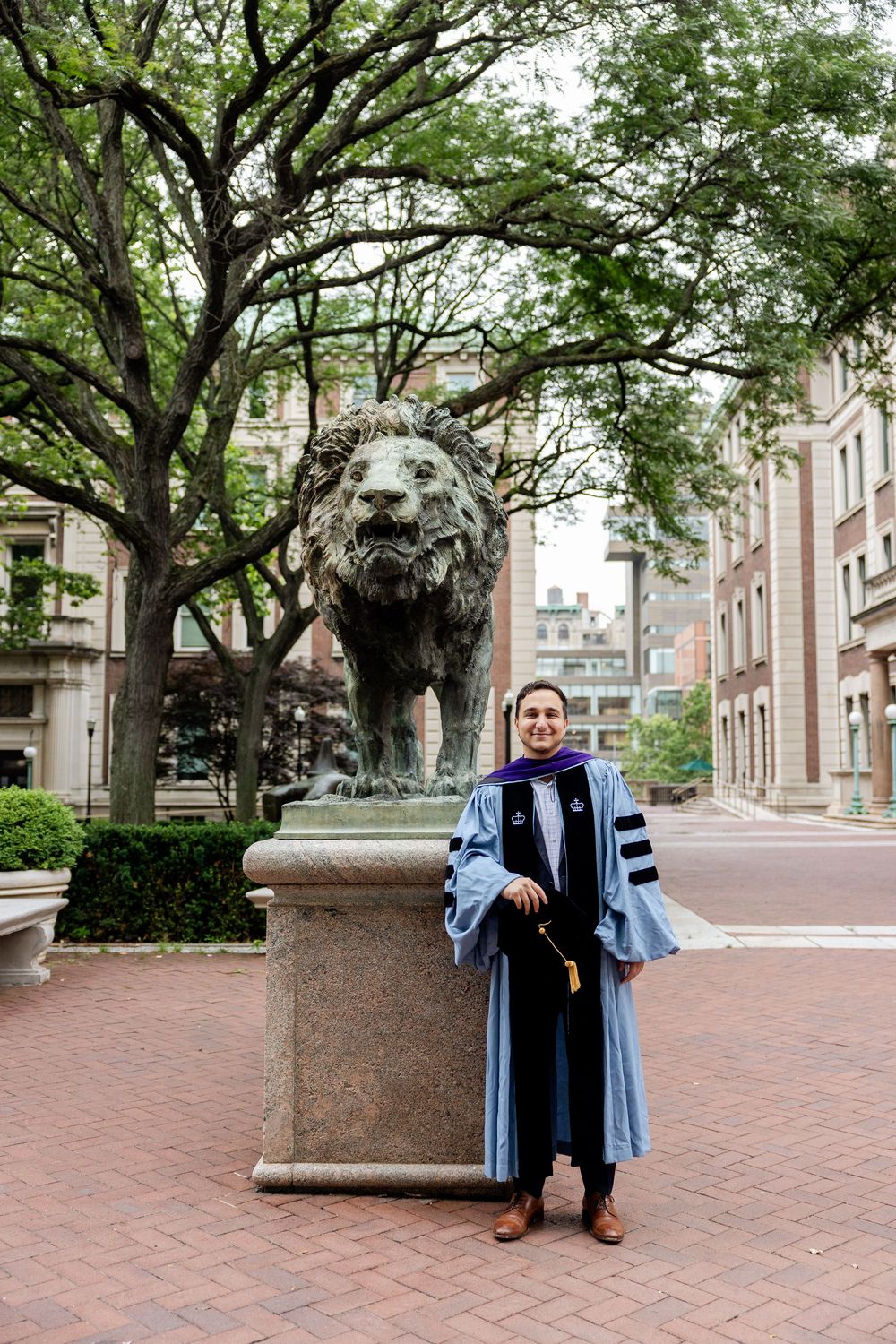 Graduate in blue academic regalia poses near brick courtyard with trees and columns.