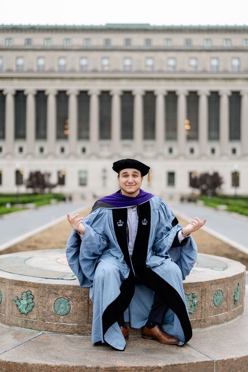 Graduate in academic regalia poses near ornate stone fountain with classical building facade.
