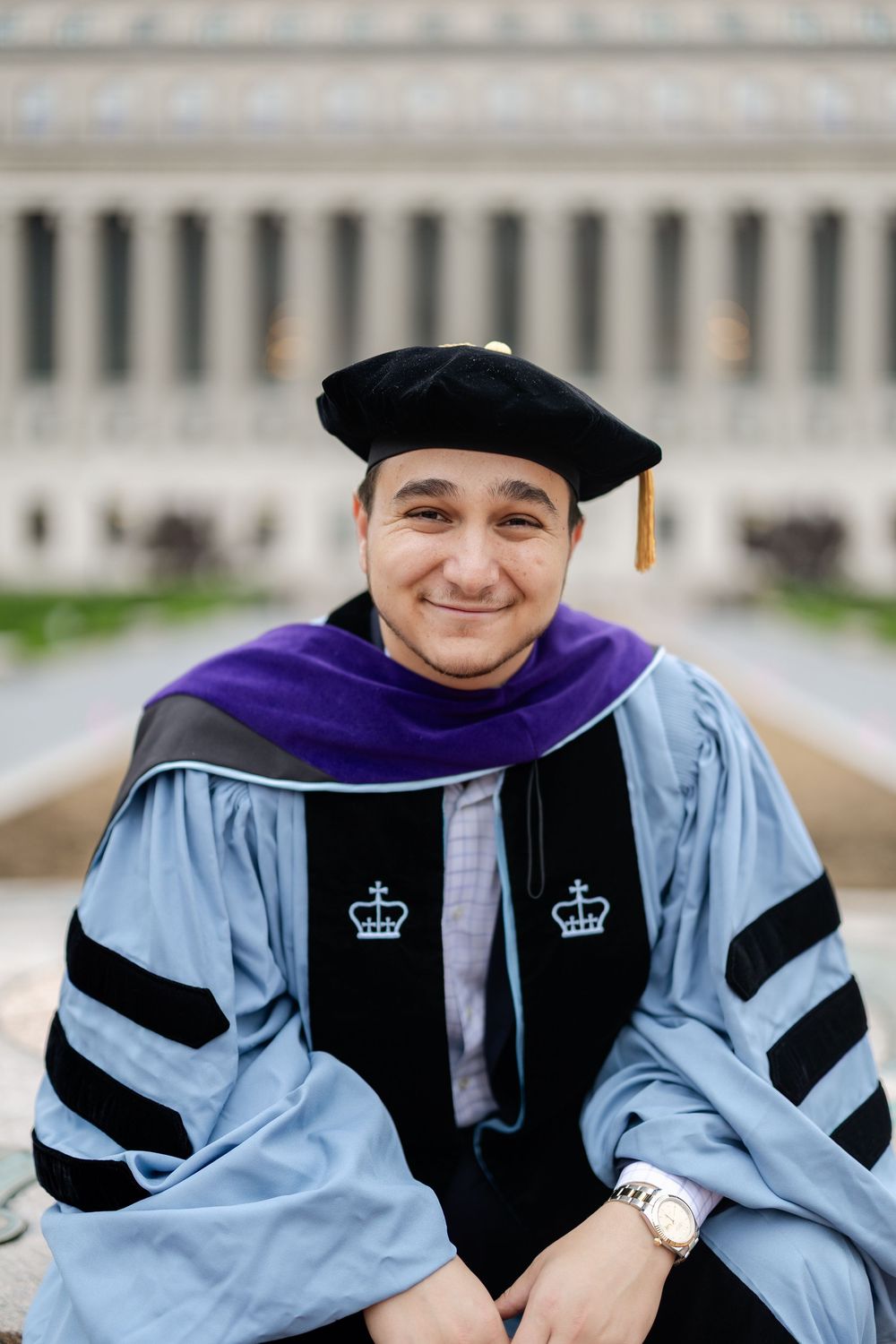 Graduate in academic regalia poses with light blue and black doctoral robes and purple hood against architectural backdrop.