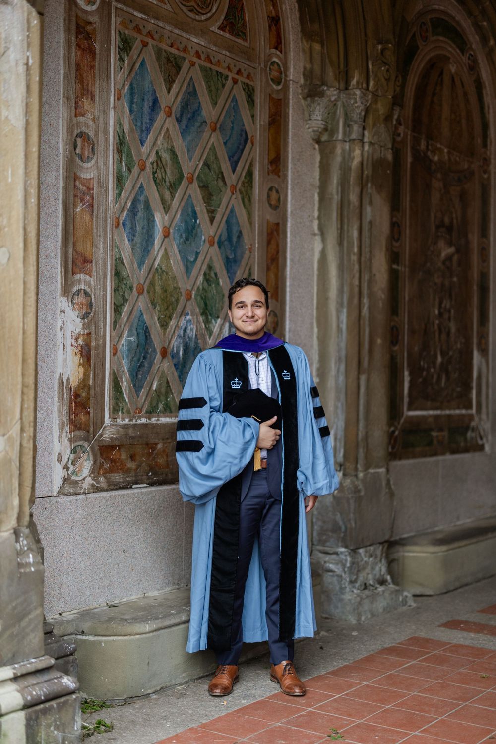 Academic regalia against ornate painted archways and terracotta tiles in historic corridor.