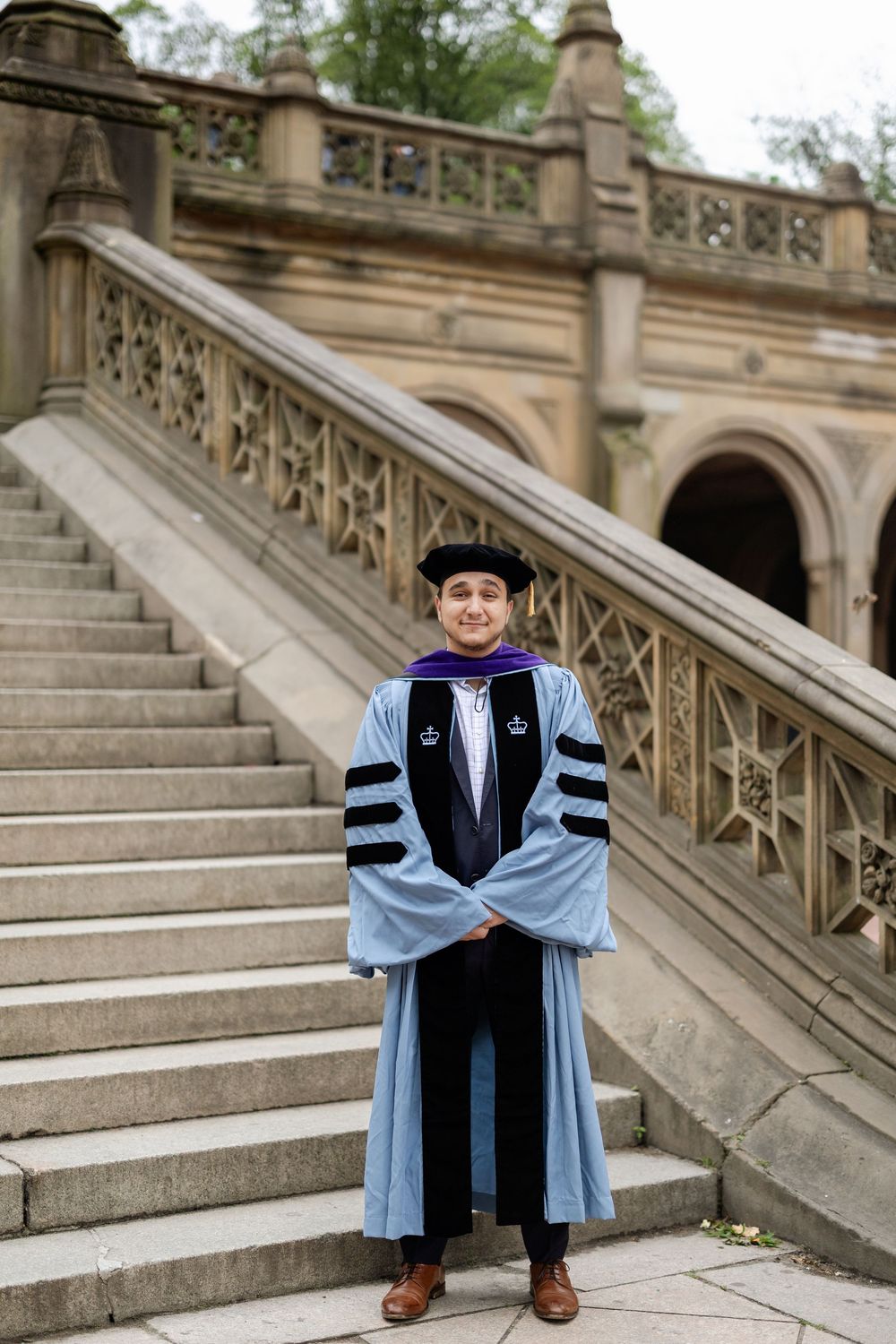 Academic graduate sits on ornate stone steps wearing doctoral regalia with light blue and black robes.