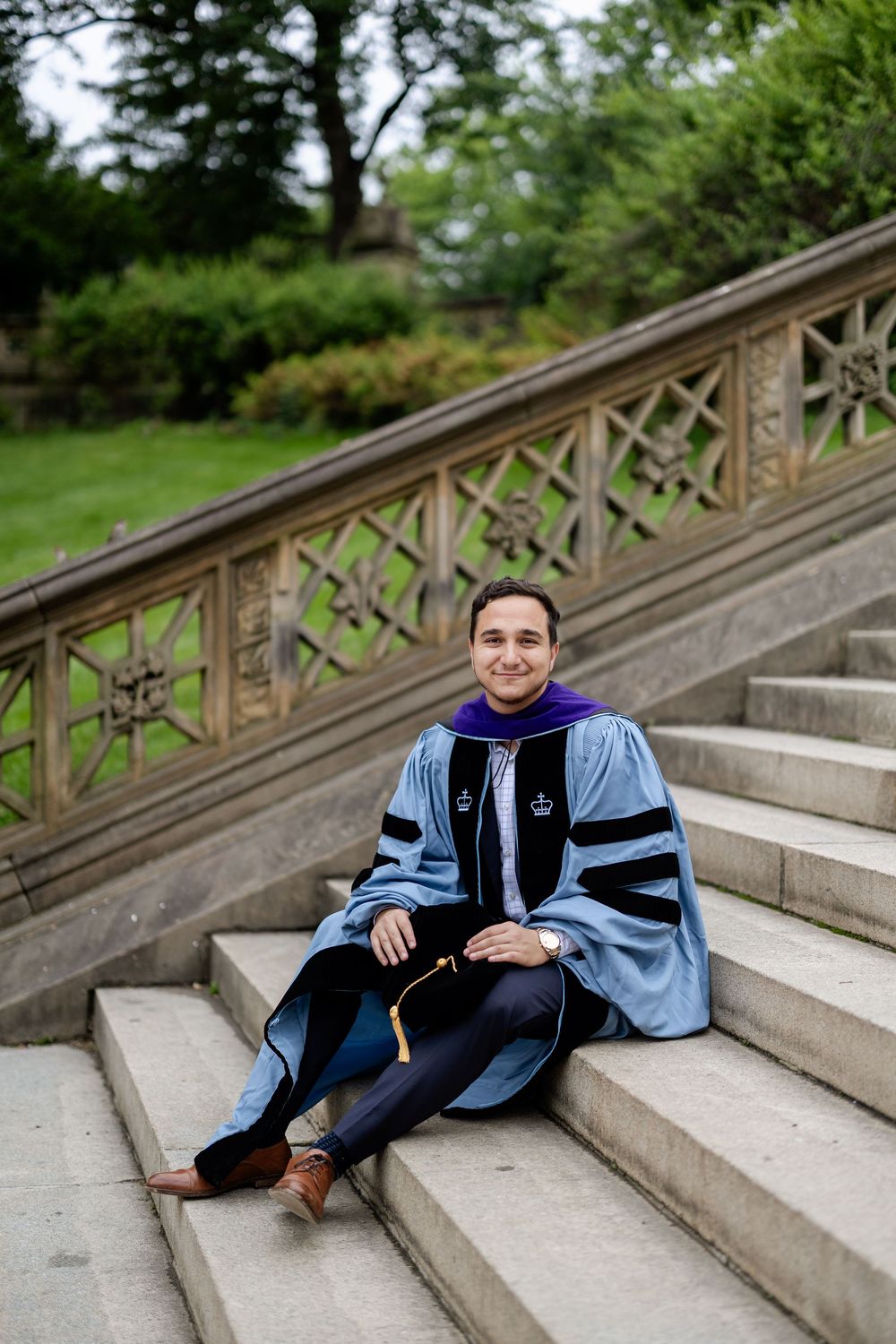 Graduate in academic regalia reclines on stone steps surrounded by greenery.