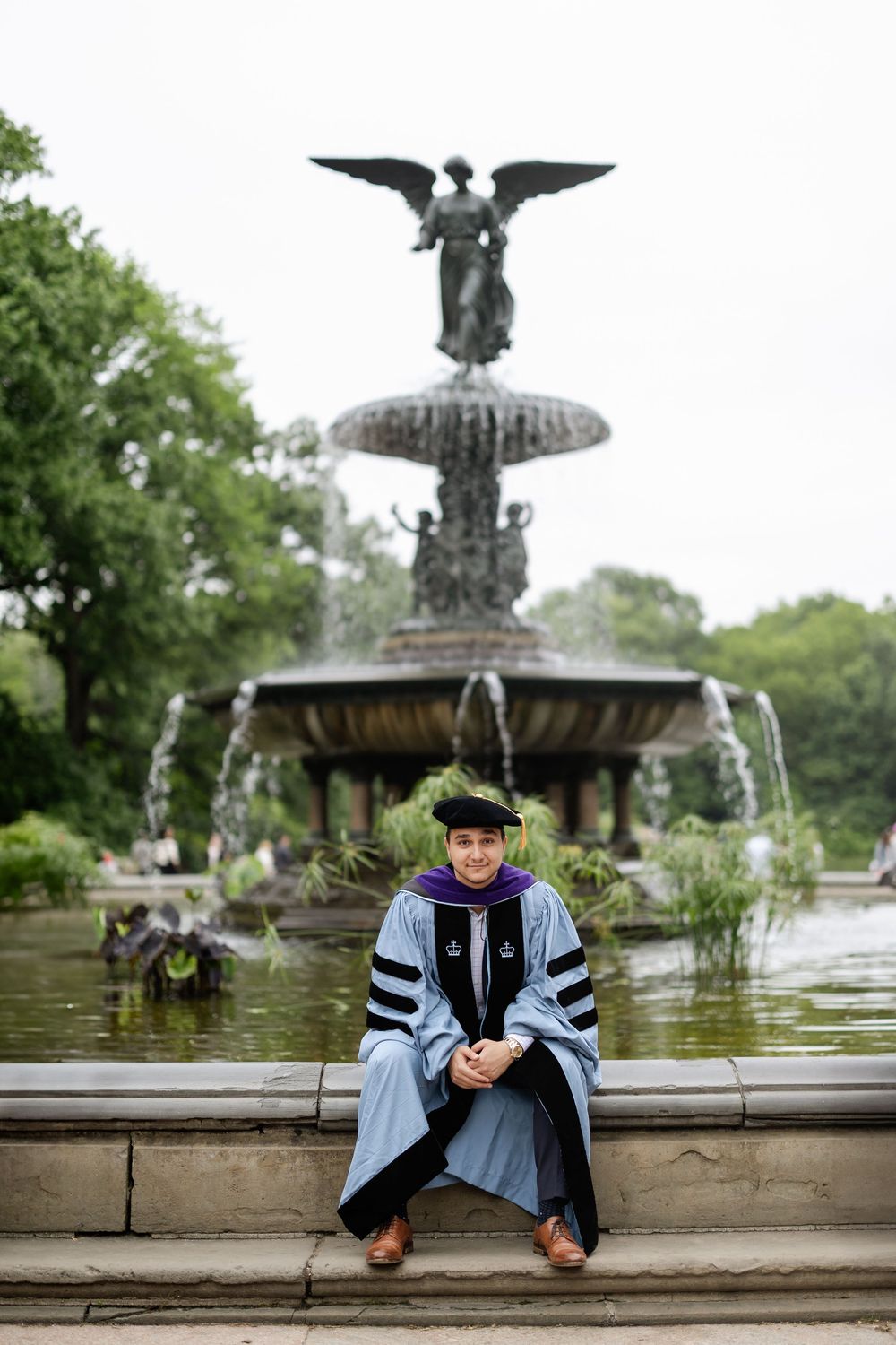 Graduate in academic dress sits by historic fountain with eagle statue in background.