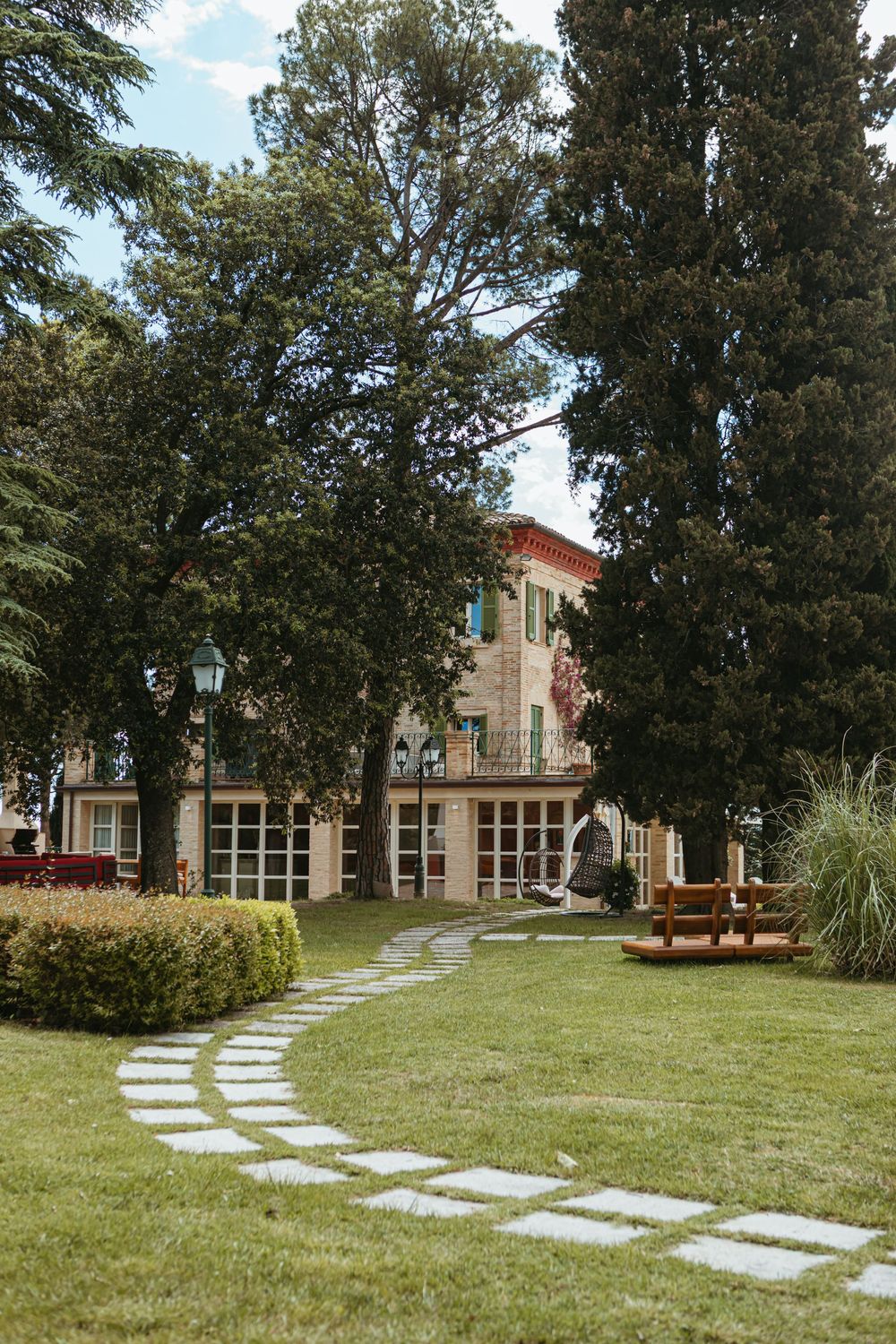 Winding stone path leads through lush green garden to elegant Italian villa surrounded by tall trees.