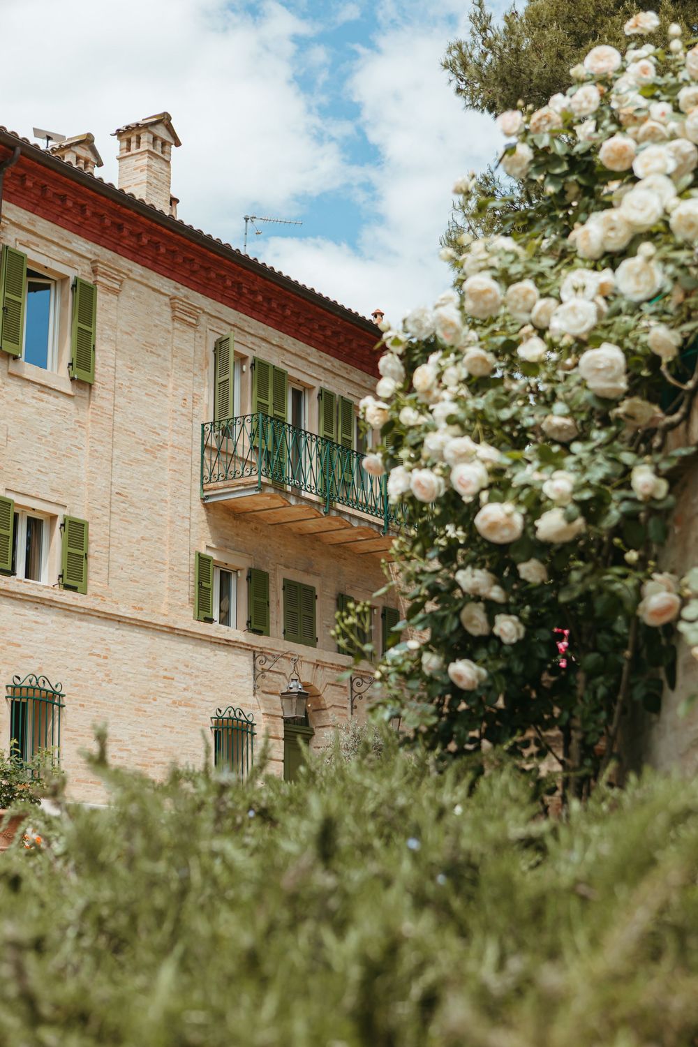 Charming Italian stone villa with green shutters and balcony, framed by blooming white roses under a blue sky.
