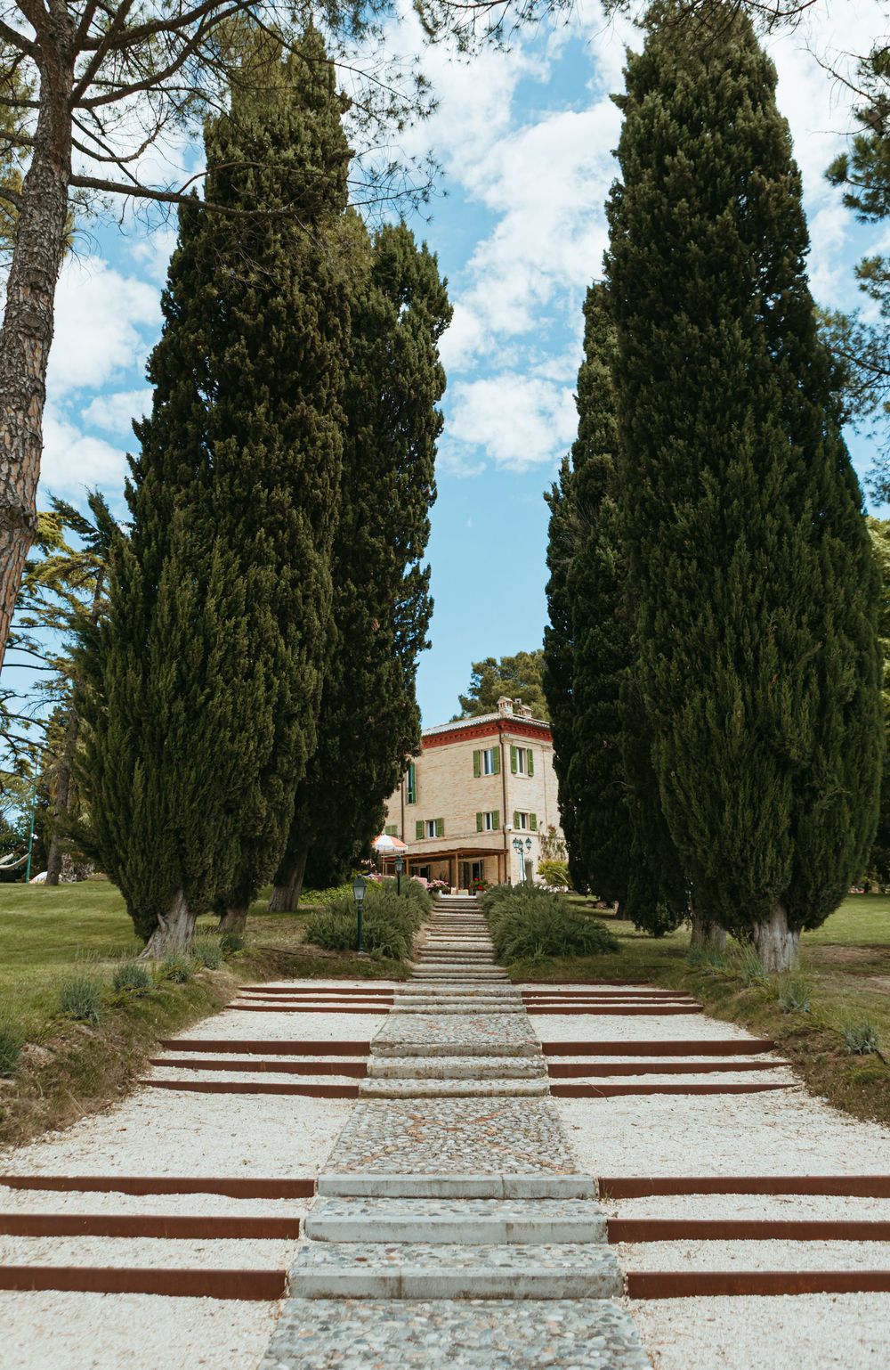 Grand cypress-lined stone pathway leads to a classic Italian villa under a bright blue sky.