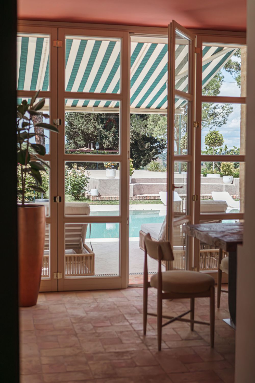 Elegant interior with French doors opening to a sunlit pool terrace, striped green awning, and lush garden views.