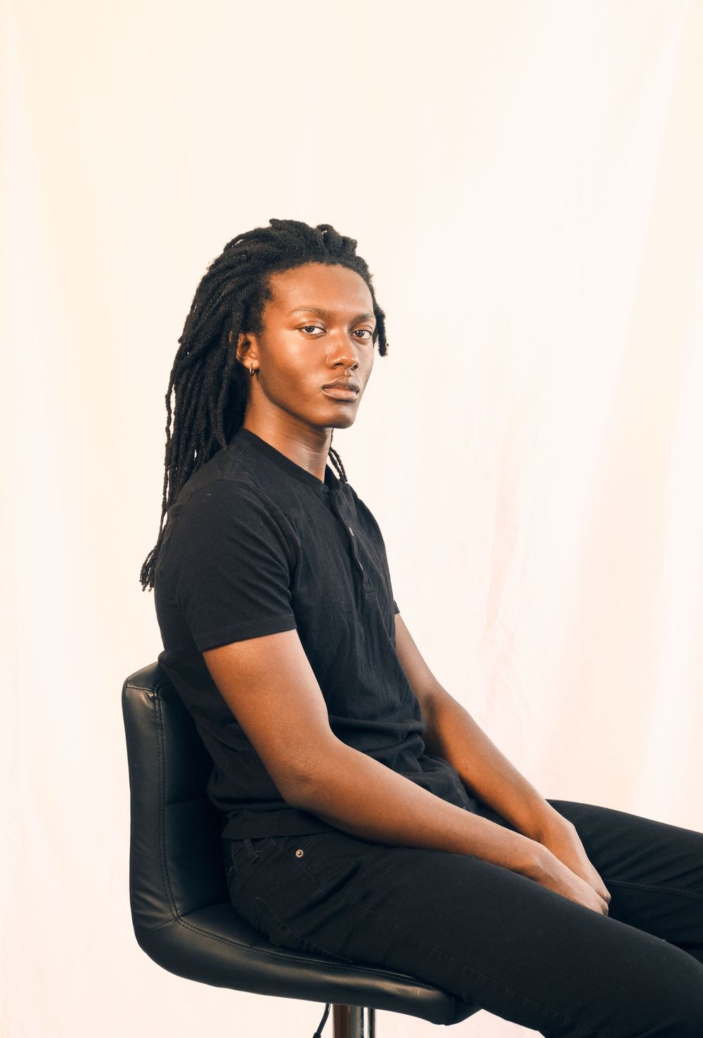 A person with dreadlocks wearing a black shirt sits in profile view on a chair against a white background.