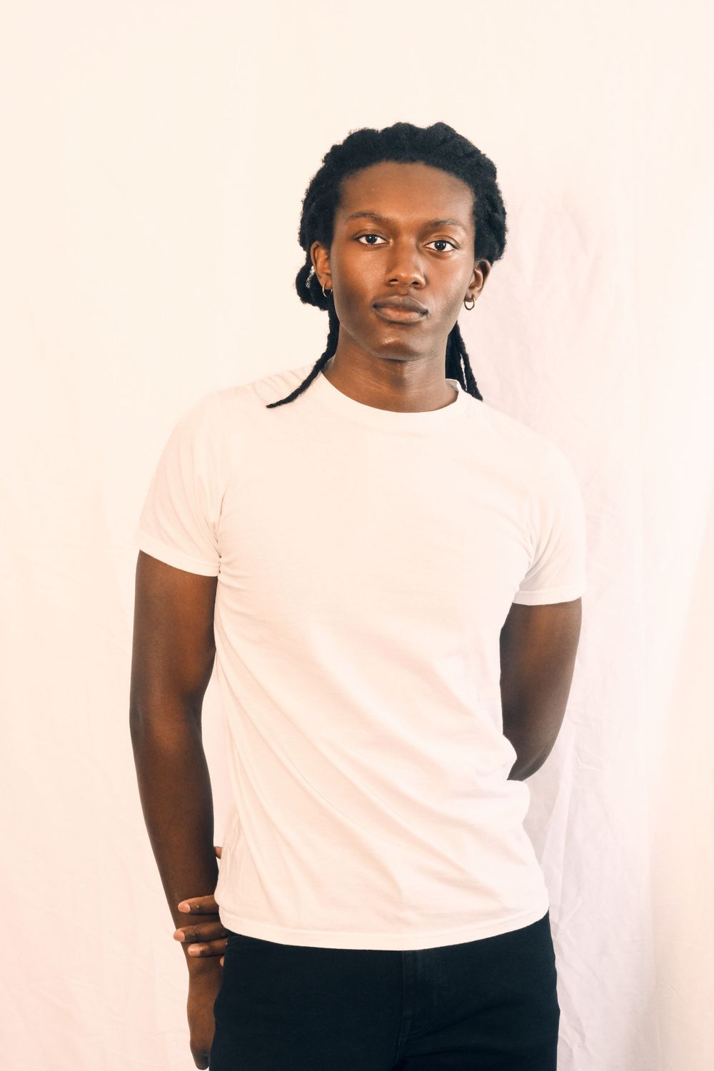A person in a white t-shirt and dark pants poses against a light colored wall.