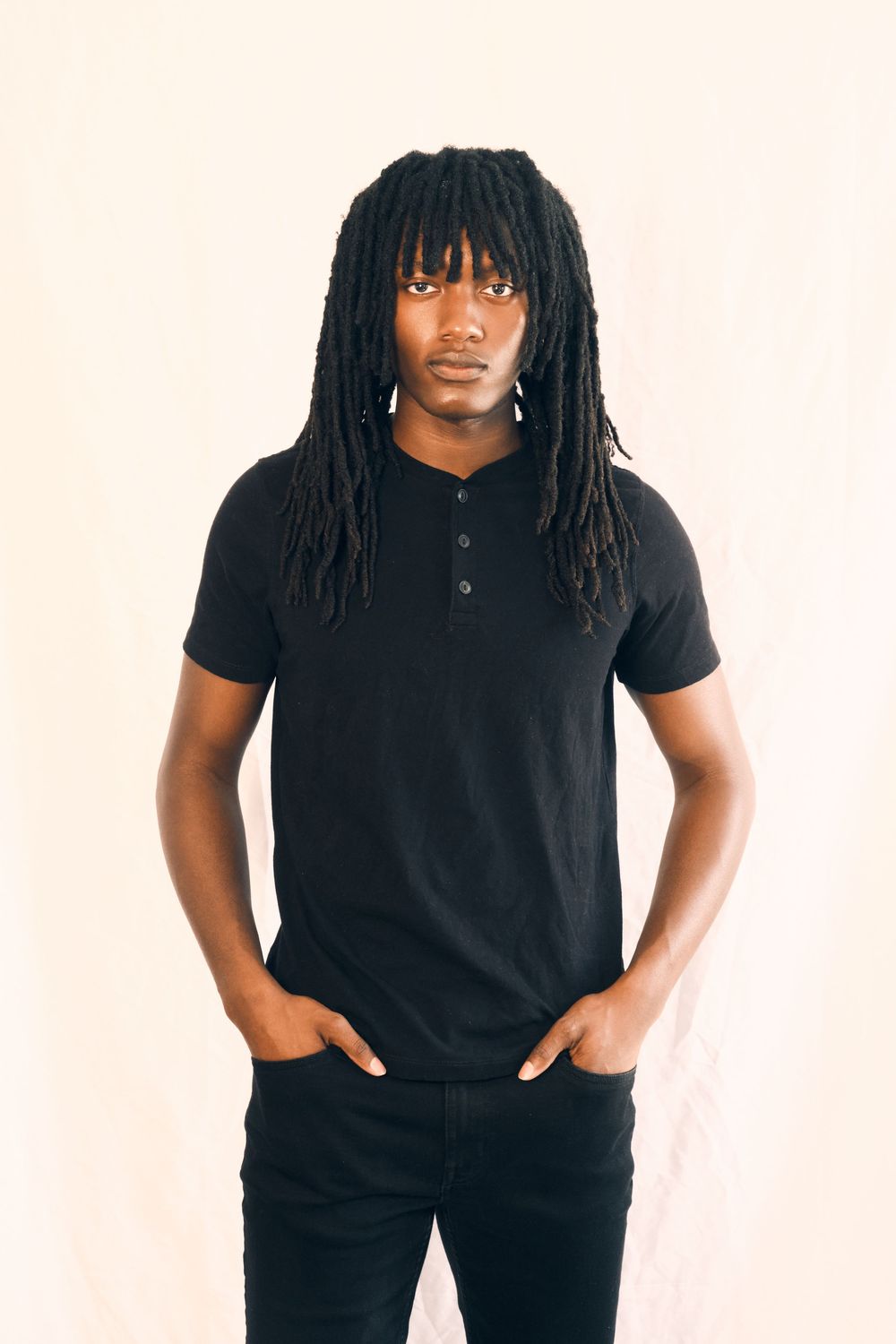 Portrait of someone wearing a black henley shirt and dark pants with long dreadlocks against a light background.