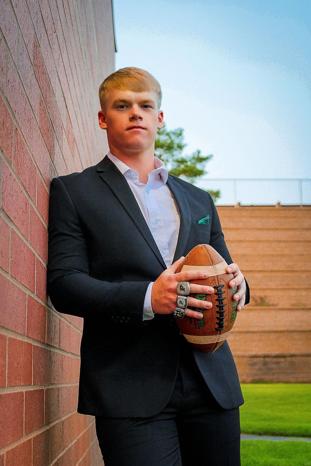 Young athlete in dark suit poses against brick wall holding football with confident stance and formal attire.