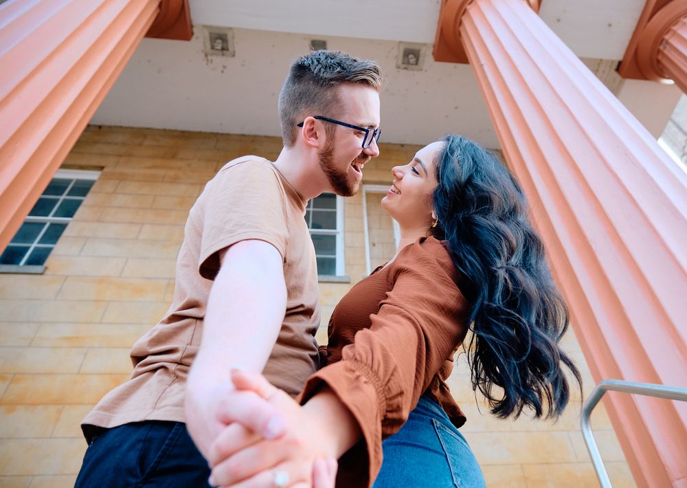 A couple embraces and dances together in front of pink columns of a classical building exterior.