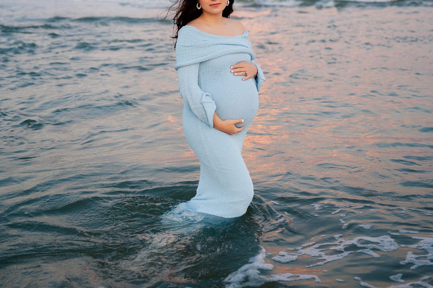 A maternity photoshoot at the beach with flowing blue dress and ocean waves at sunset.