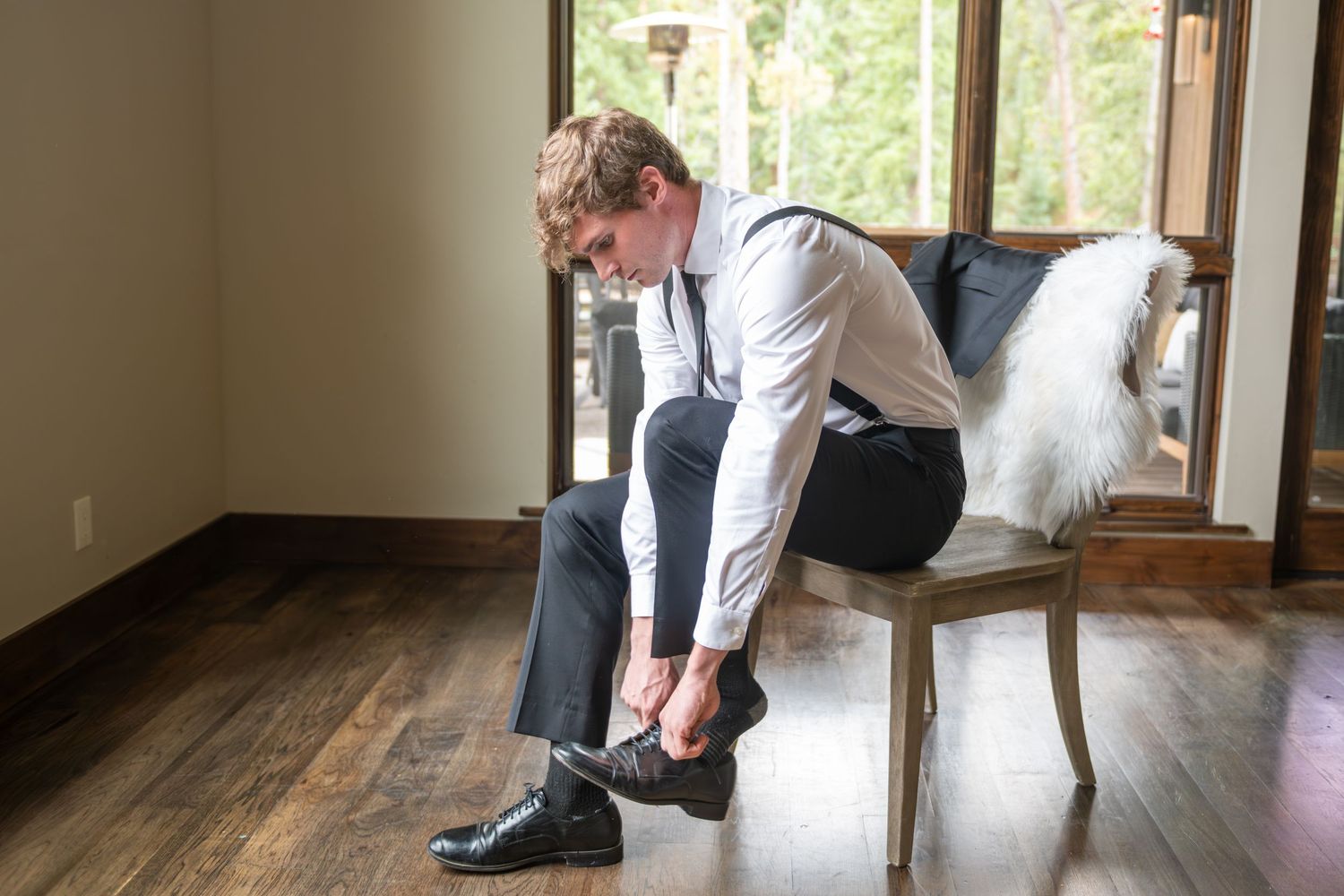 A groom is sitting in a brown chair in front of a window putting on his shoes. He is in natural window light.