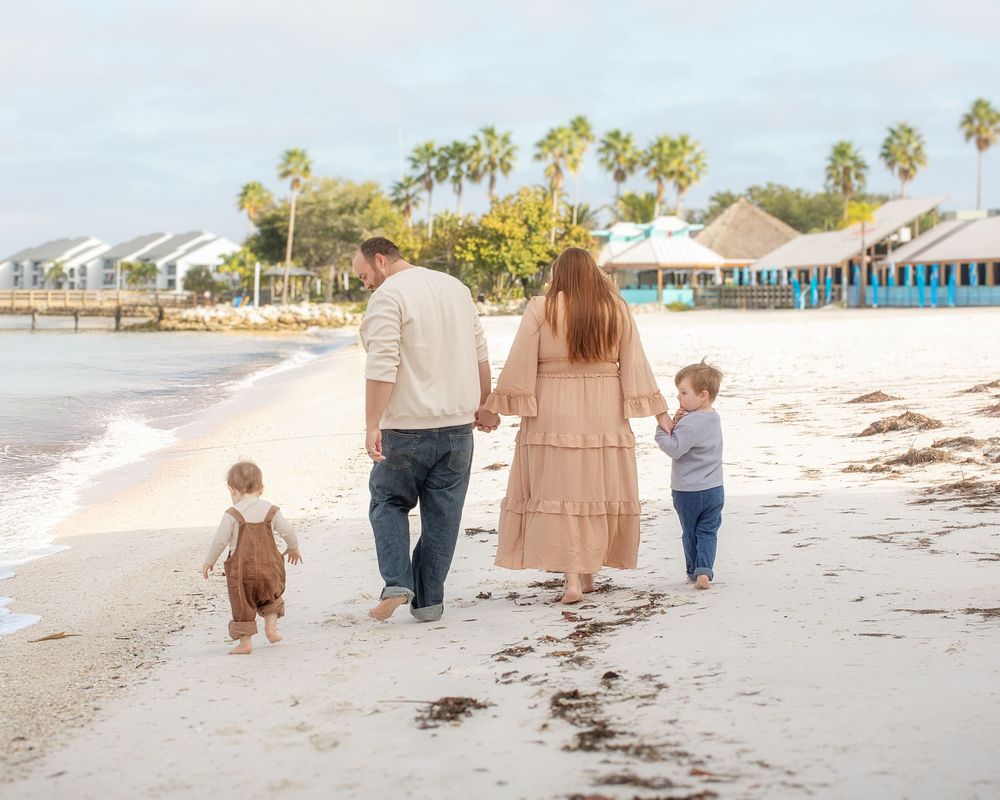 A family walks together along a serene beach with palm trees and a pier in the distance.