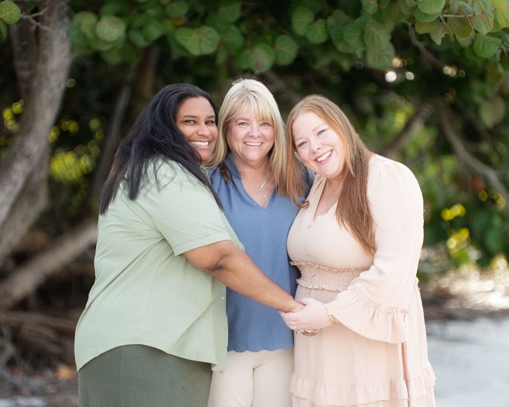 Three people embrace and smile together outdoors on a sunny day with greenery in the background.