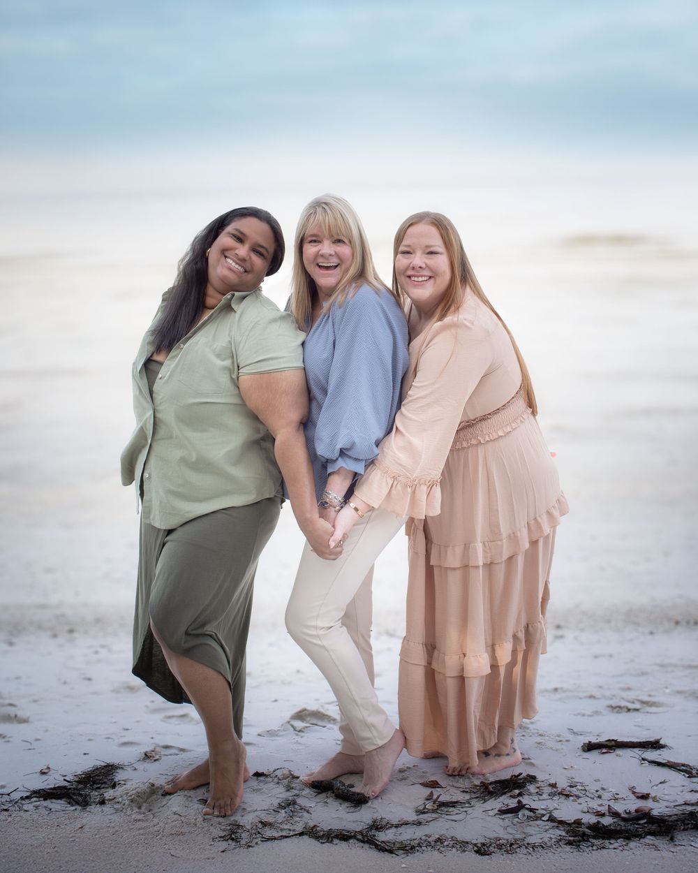 Three friends in pastel dresses pose playfully together on a sandy beach at sunset.