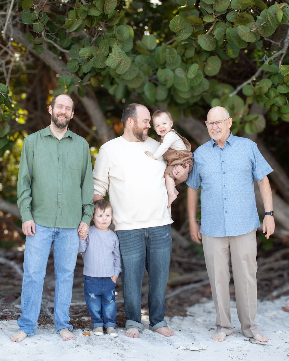 Family group poses together on sandy beach with green foliage in background.