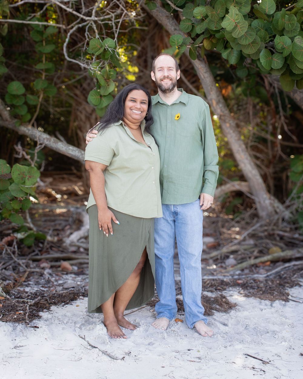 Two people stand together on white sandy beach surrounded by lush green foliage.