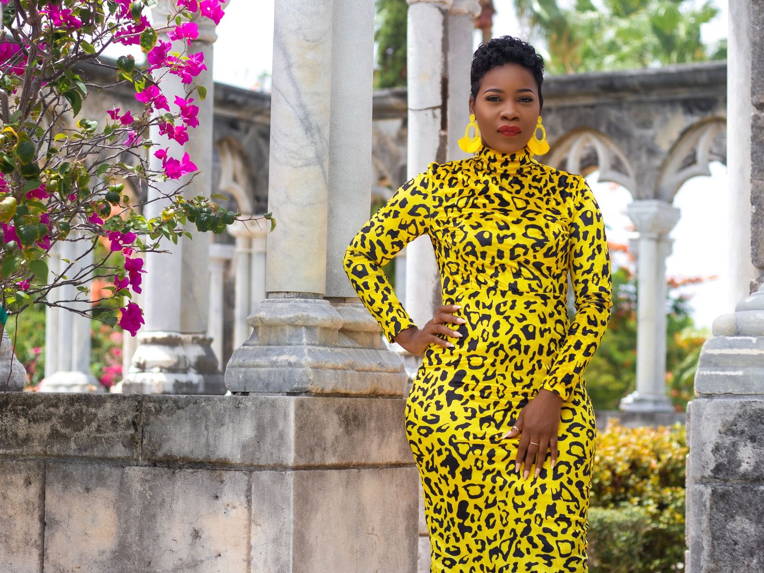 A person wearing a vibrant yellow leopard print outfit poses among pink flowers and white columns.