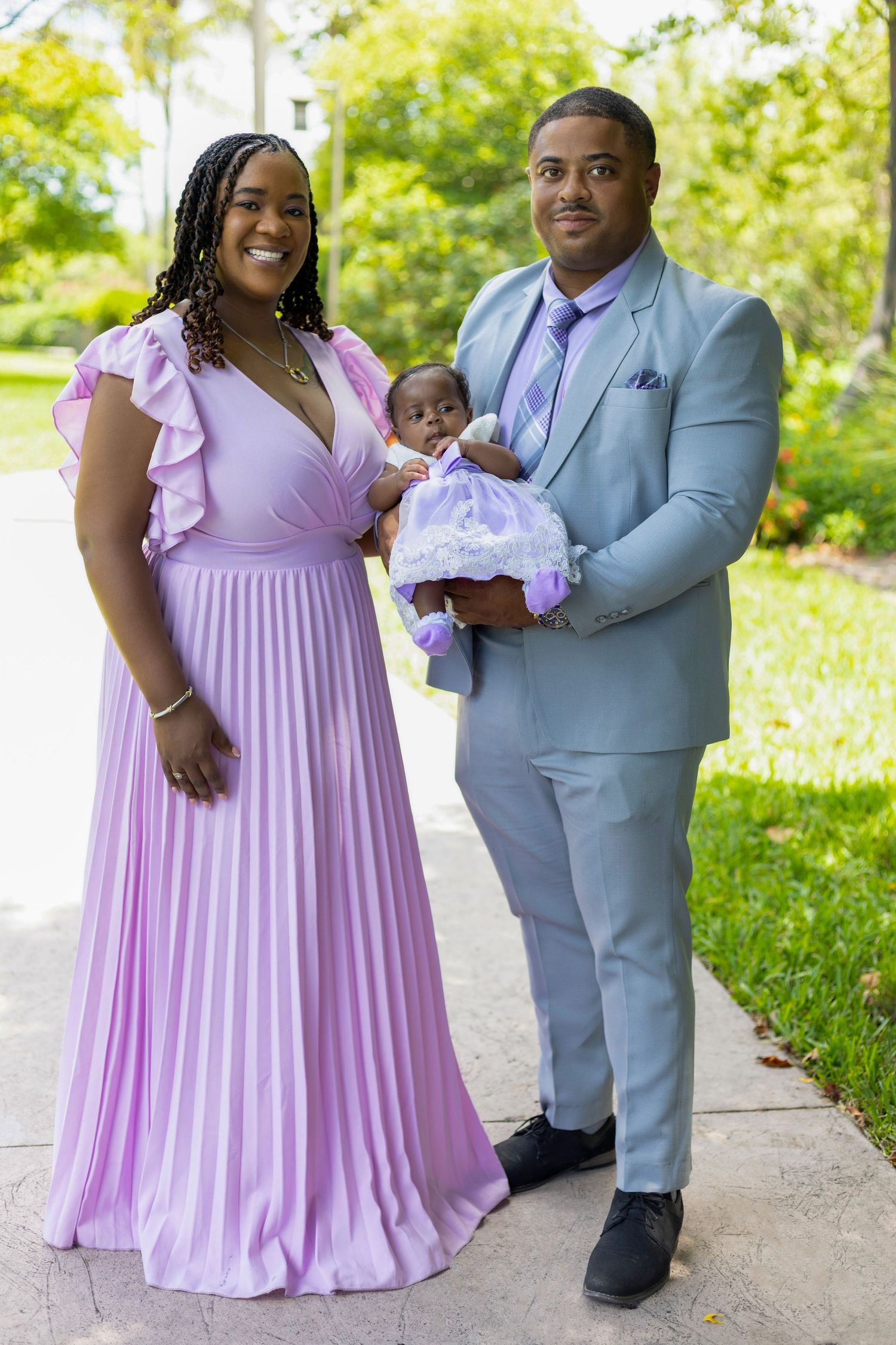 A couple in coordinating lavender and gray formal attire pose outdoors with their baby in a garden setting.
