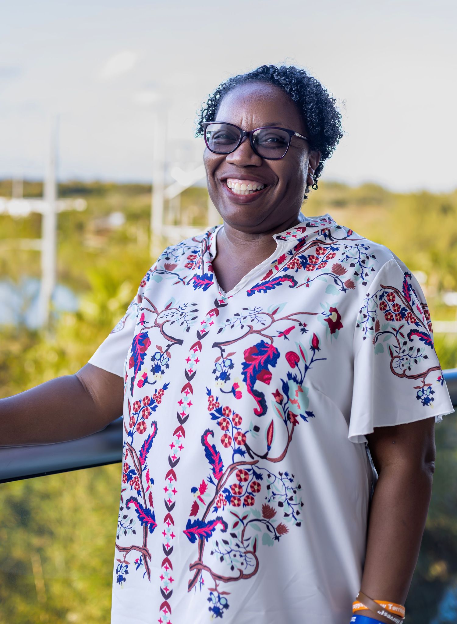 Smiling woman in floral print blouse wearing glasses, standing on outdoor balcony with green landscape background.