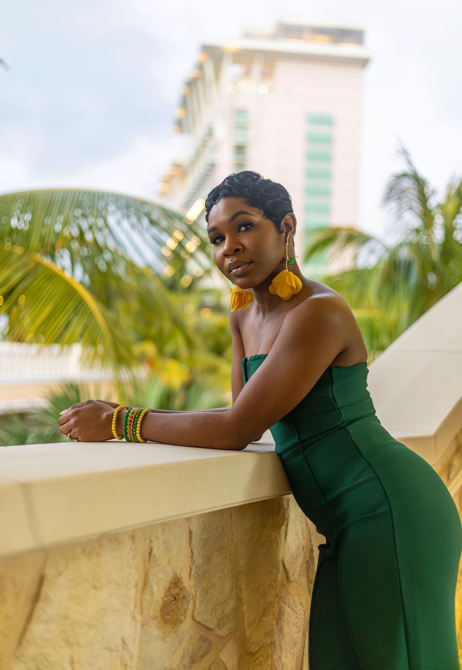 A woman in a green strapless dress with yellow earrings and bracelets poses on a tropical resort balcony.