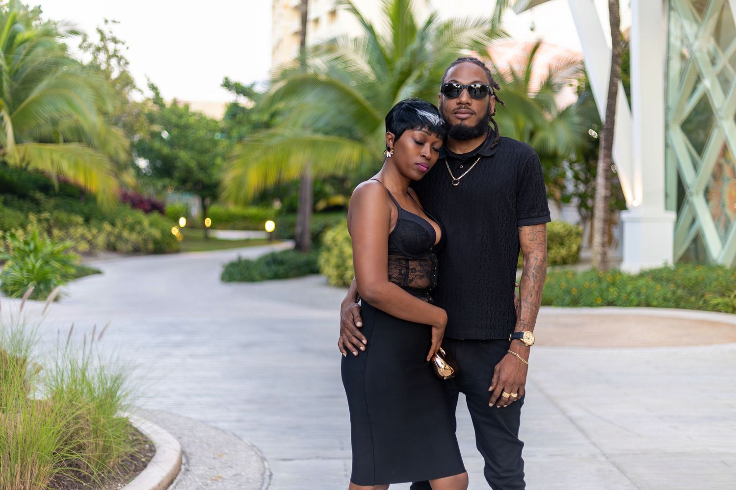 A couple in matching black outfits poses together on a tropical resort pathway surrounded by lush greenery.
