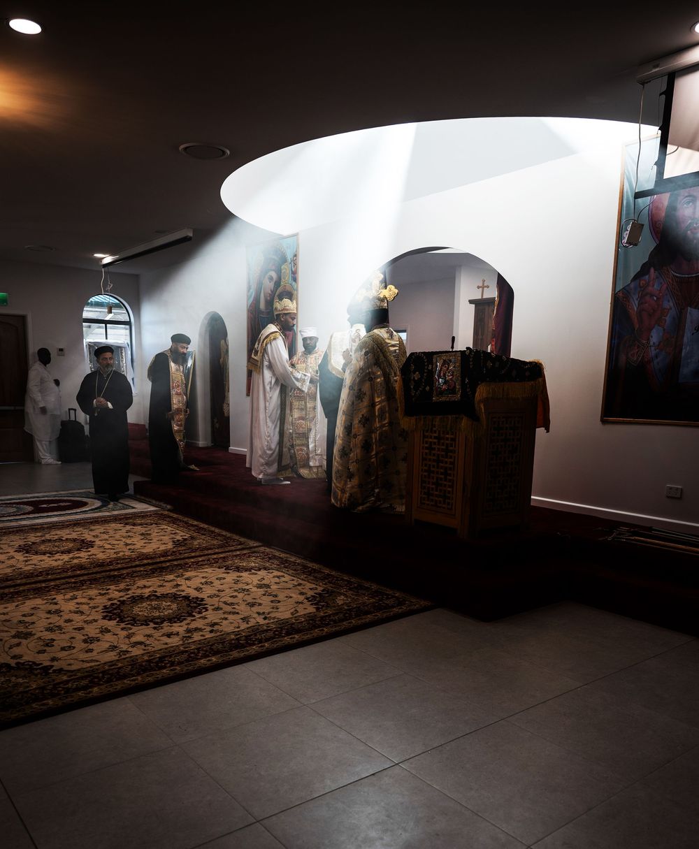 Ethiopian Orthodox priest in ornate vestments standing in dimly lit church sanctuary with dramatic light.