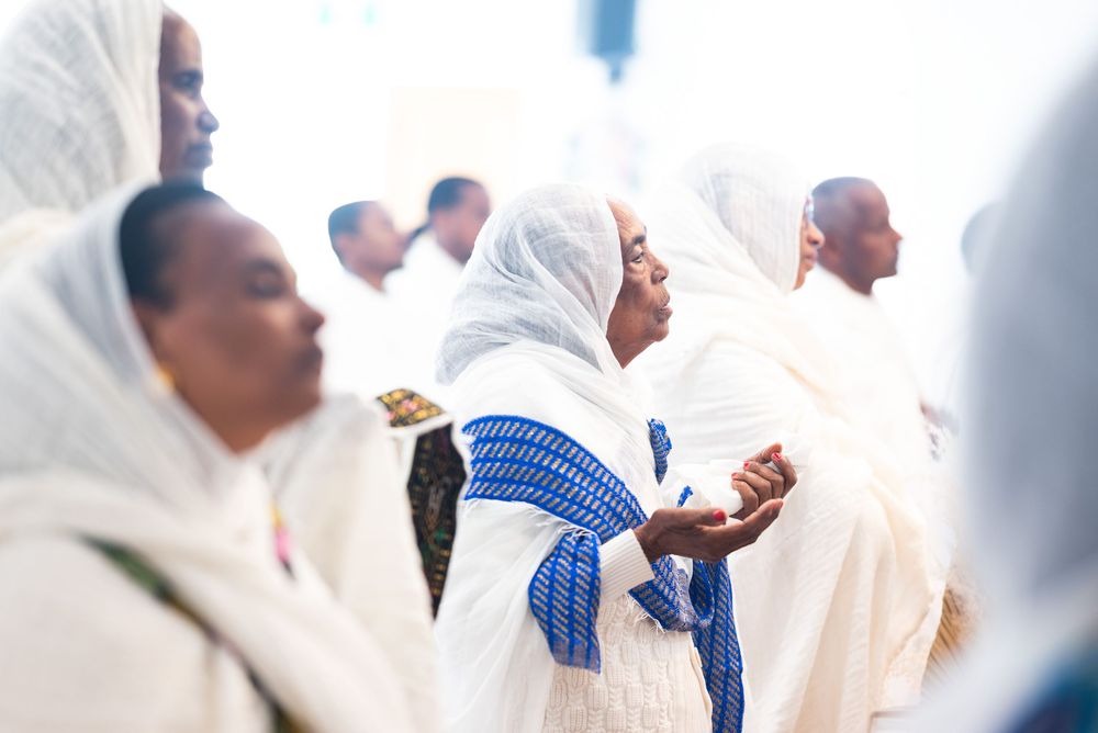 Ethiopian women in traditional white netela shawls with blue borders praying together at a religious ceremony.
