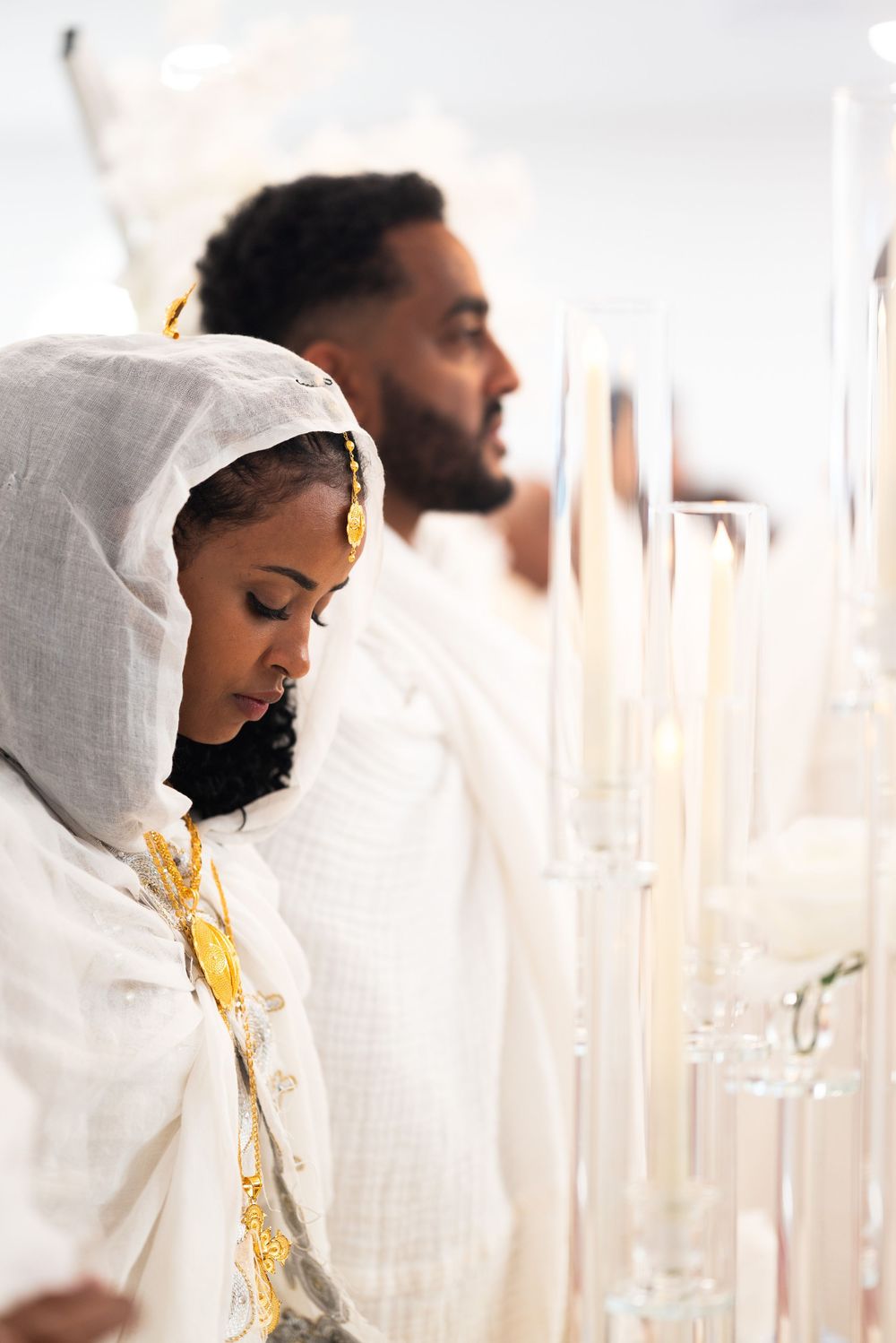 Ethiopian bride in white netela with gold accessories bowing in prayer beside candles at wedding ceremony.