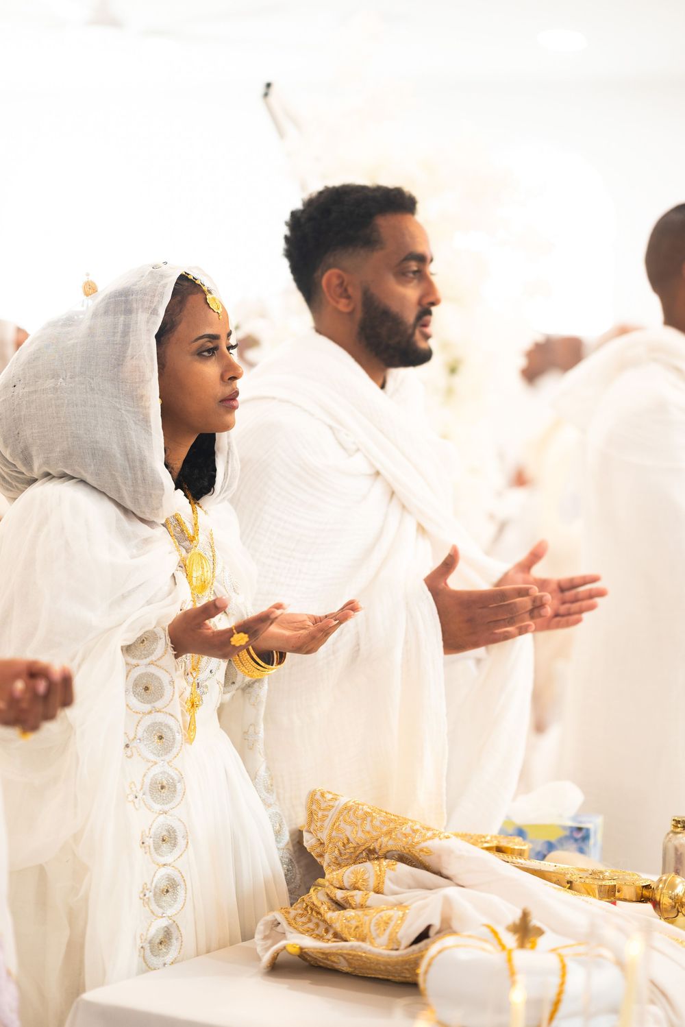 Ethiopian bride and groom in traditional white attire with gold jewelry praying with open hands at altar.