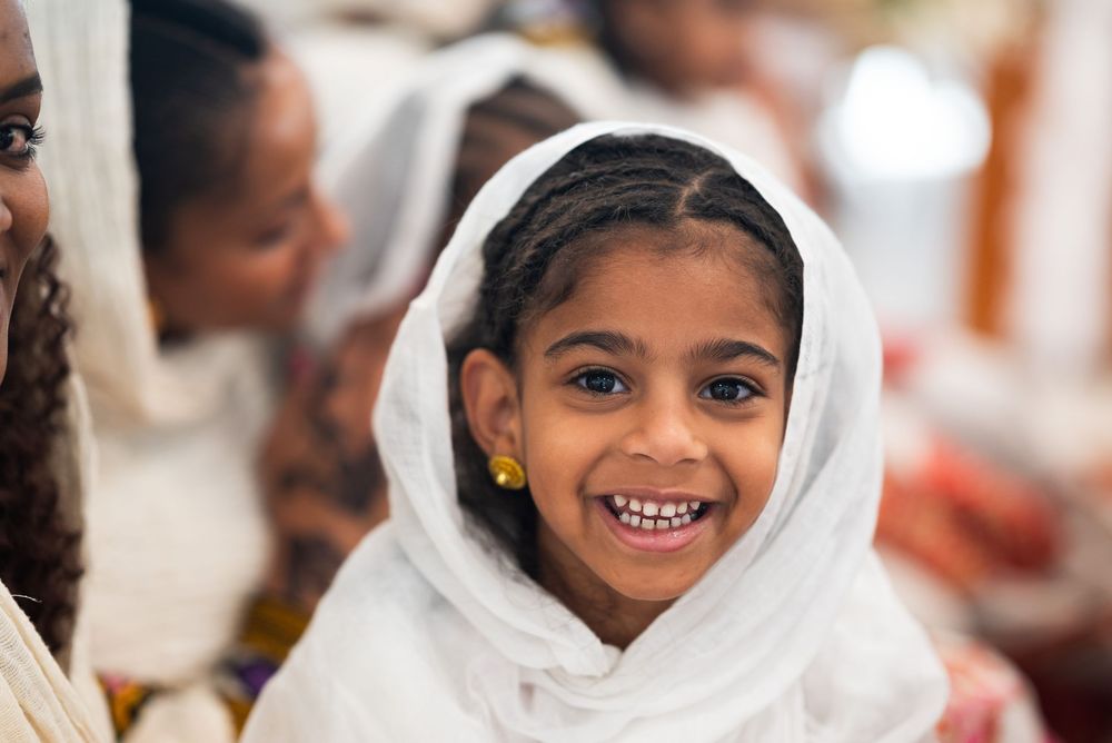 Smiling young girl wearing a white headscarf attending a joyful religious ceremony or cultural celebration.