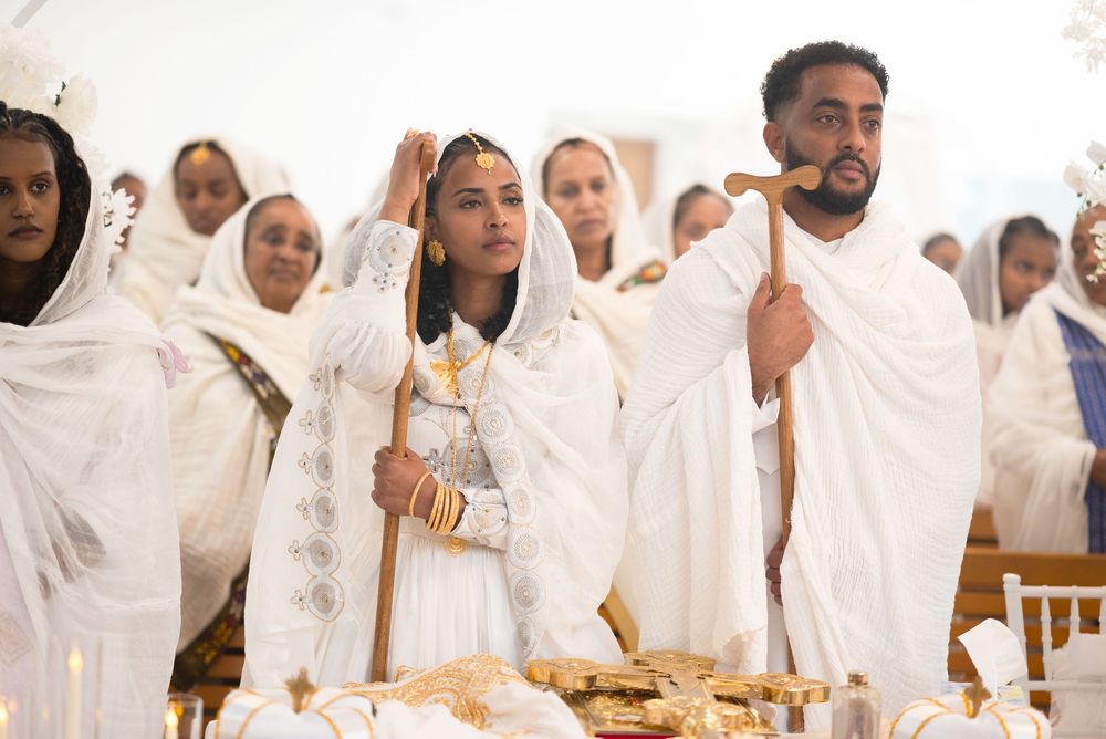 Ethiopian Orthodox wedding couple in white traditional dress holding ceremonial staffs at church altar.