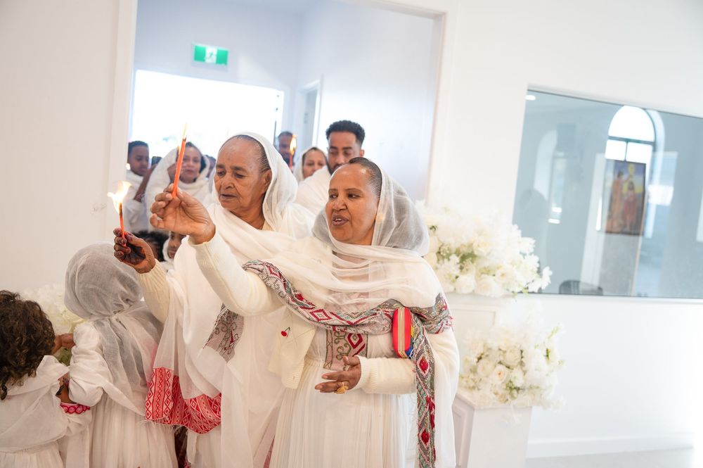 Women in white traditional Ethiopian dress with colorful trim attending a religious ceremony indoors.