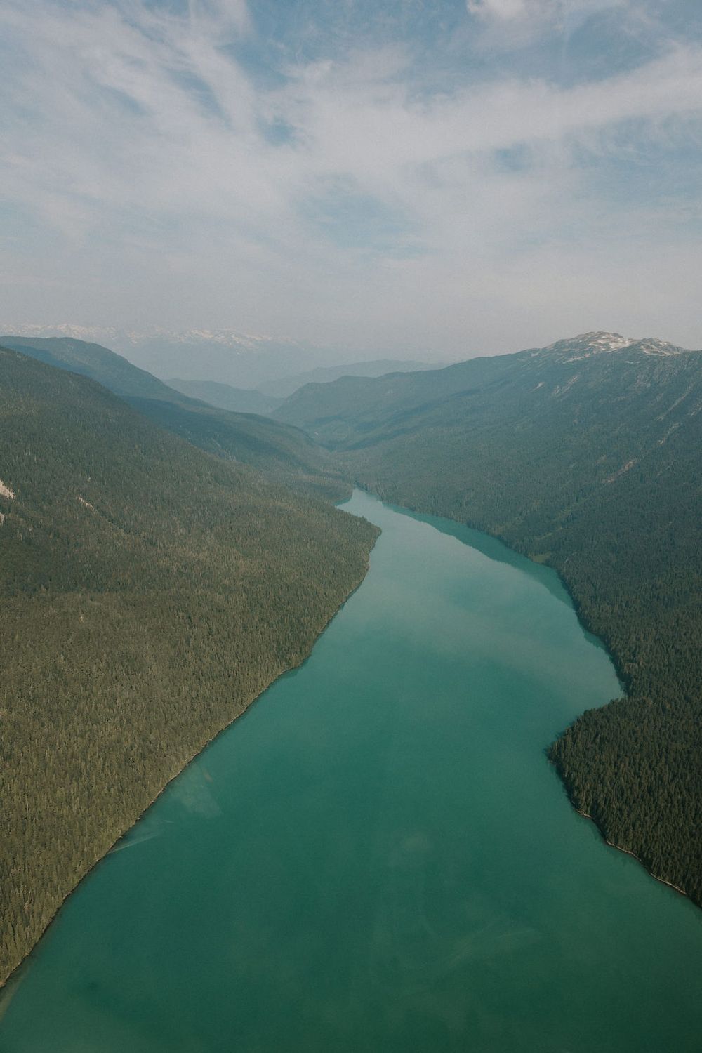 Aerial view of a winding turquoise lake surrounded by forested mountains and hills on a cloudy day.