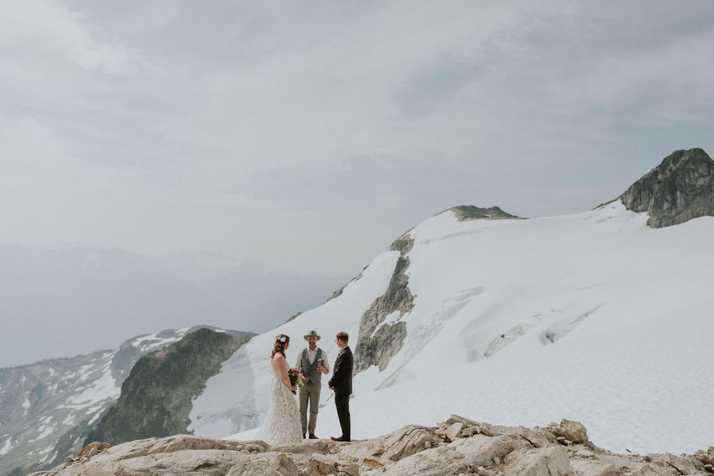 Couple in wedding attire stands on snowy mountain peak with dramatic cloudy sky and mountain ranges in background.