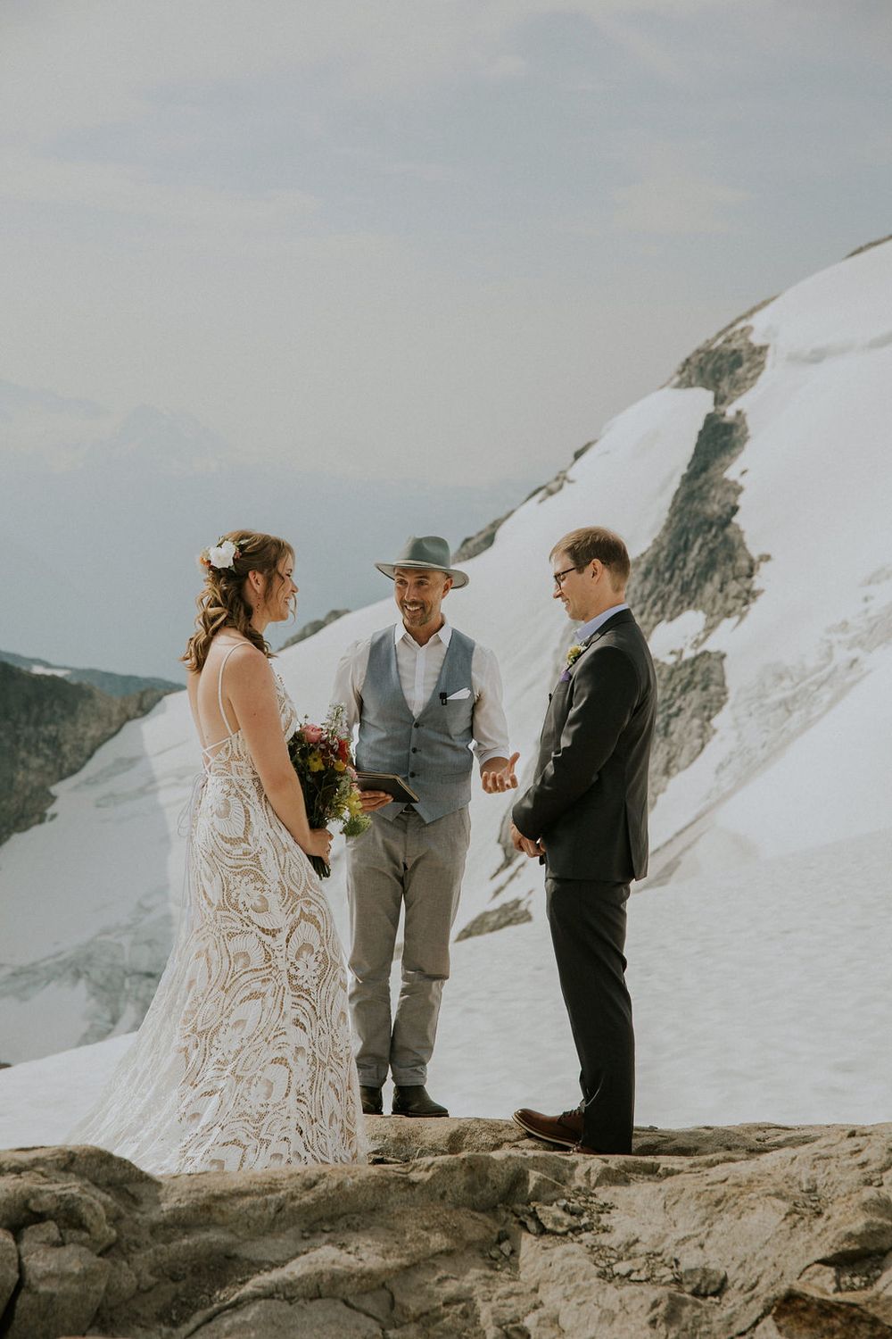 A wedding ceremony takes place on a snowy mountain peak with a couple and officiant standing on rocky terrain.