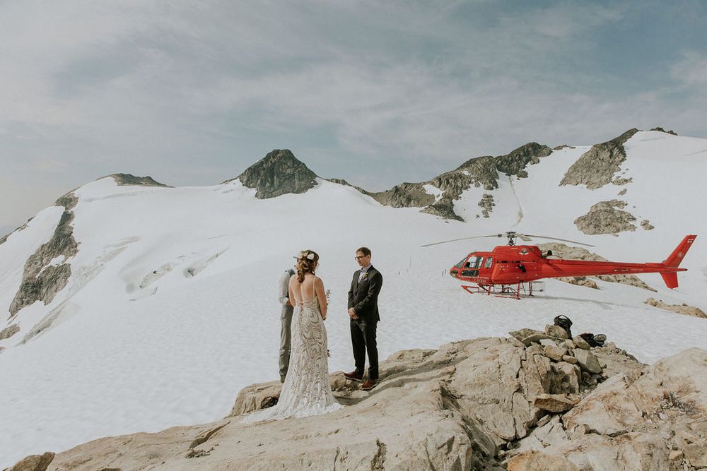 A scenic mountain wedding ceremony takes place on a snowy peak with a red helicopter in the background.