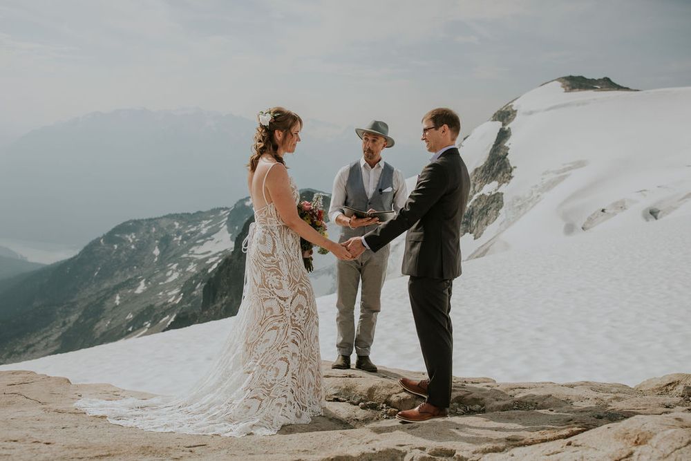 A couple exchanges vows on a dramatic mountaintop surrounded by snow and rocky terrain.