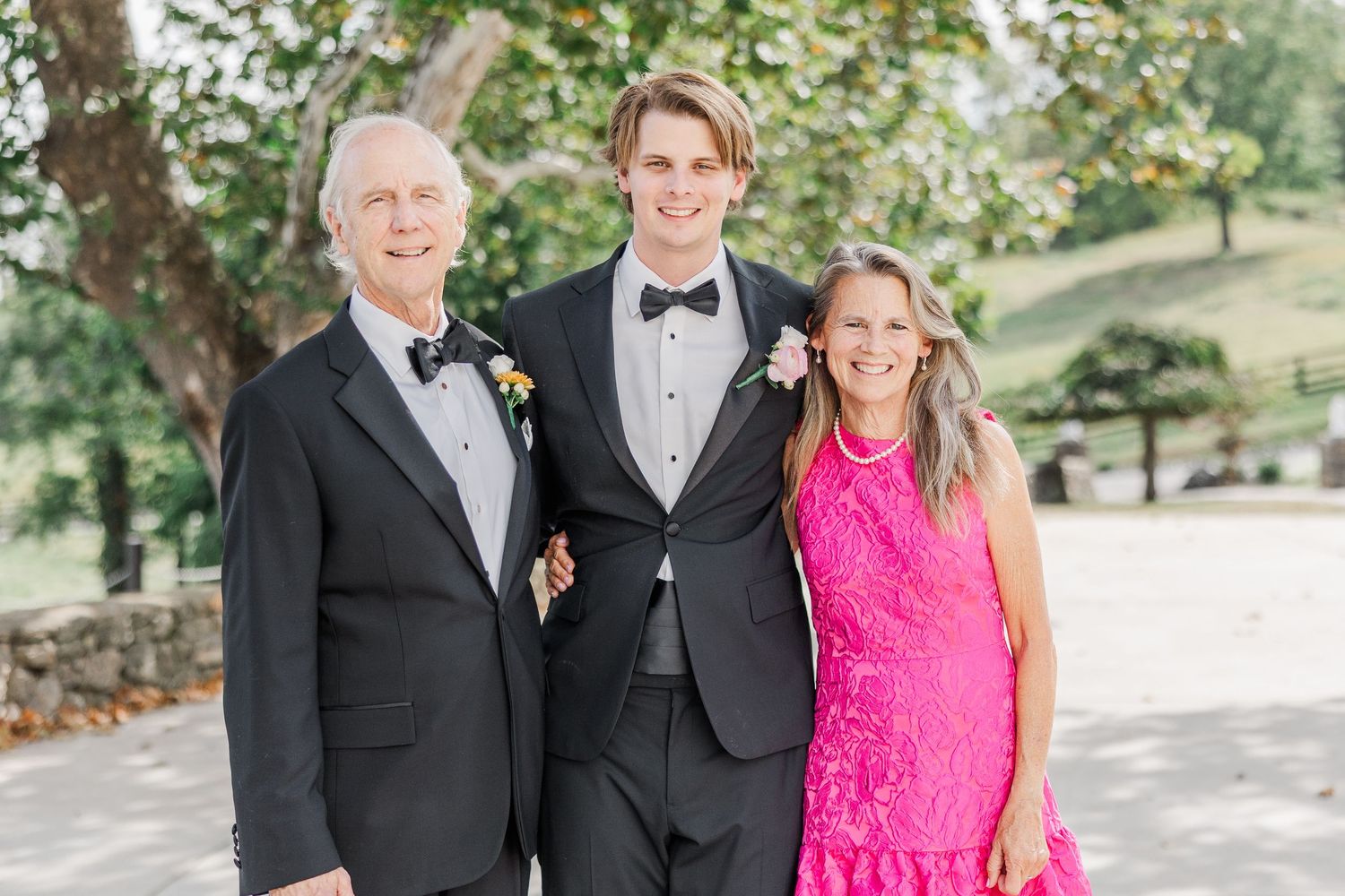 Three wedding guests in formal attire pose together outdoors with a bright pink dress adding a pop of color.