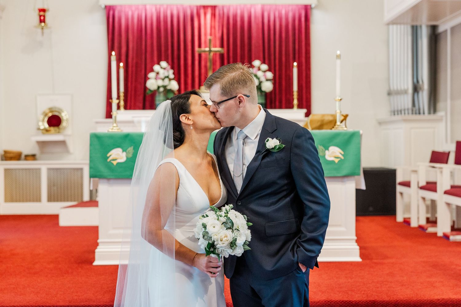 A couple shares an intimate kiss during their wedding ceremony in a church with red carpeting and white pews.