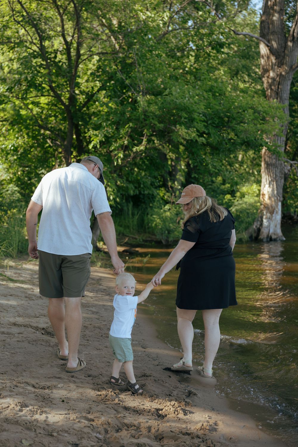 Three people walking together along a riverbank path surrounded by lush green trees in a park setting.