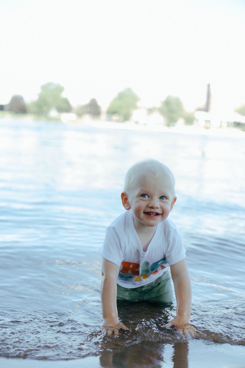 A happy toddler plays in shallow water at the edge of a lake on a bright summer day.