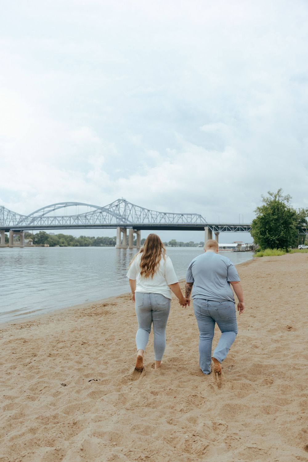 Two people in matching gray outfits walk together on a sandy beach with a bridge visible in the background.